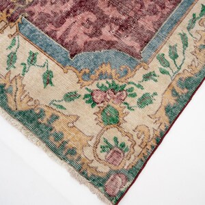 May include: Close-up of a section of a vintage rug with a distressed appearance. The rug features a central maroon field with a floral pattern, framed by a light beige border with green and gold floral motifs. The edges are fringed.