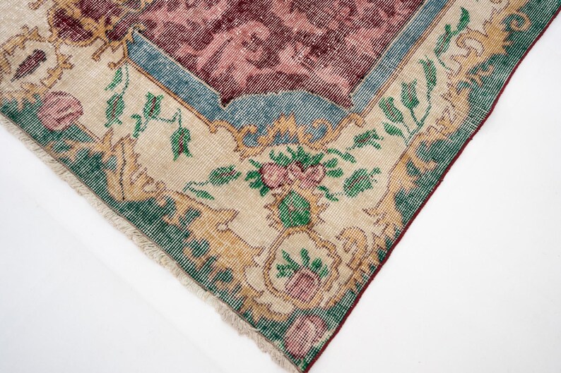 May include: Close-up of a section of a vintage rug with a distressed appearance. The rug features a central maroon field with a floral pattern, framed by a light beige border with green and gold floral motifs. The edges are fringed.