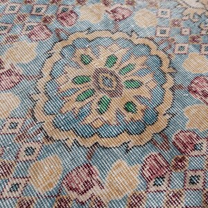 May include: Close-up of a vintage rug with a central floral medallion in shades of blue, beige, and green. The rug features a repeating geometric pattern and accents of pink and burgundy, creating a textured, antique appearance. The rug is likely made of wool.