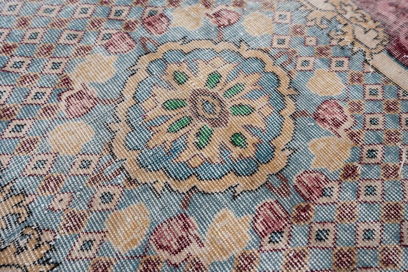 May include: Close-up of a vintage rug with a central floral medallion in shades of blue, beige, and green. The rug features a repeating geometric pattern and accents of pink and burgundy, creating a textured, antique appearance. The rug is likely made of wool.