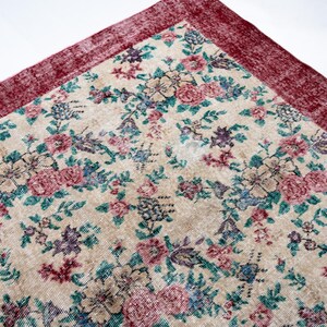 Pode incluir: Um canto de um tapete com padr&atilde;o floral com fundo creme. O tapete apresenta um padr&atilde;o repetido de flores rosa, azuis e roxas com folhas verdes. A borda &eacute; de cor bord&ocirc; profundo.