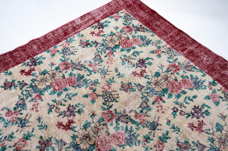 Pode incluir: Um canto de um tapete com padr&atilde;o floral com fundo creme. O tapete apresenta um padr&atilde;o repetido de flores rosa, azuis e roxas com folhas verdes. A borda &eacute; de cor bord&ocirc; profundo.
