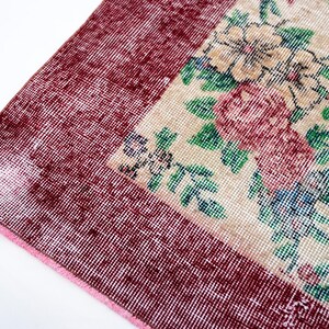 Pode incluir: Close-up de um tapete vintage com um padr&atilde;o floral. O tapete apresenta uma borda bord&ocirc; e um design central com flores rosa, verdes e azuis sobre um fundo bege. O tapete tem uma apar&ecirc;ncia desgastada e antiga.