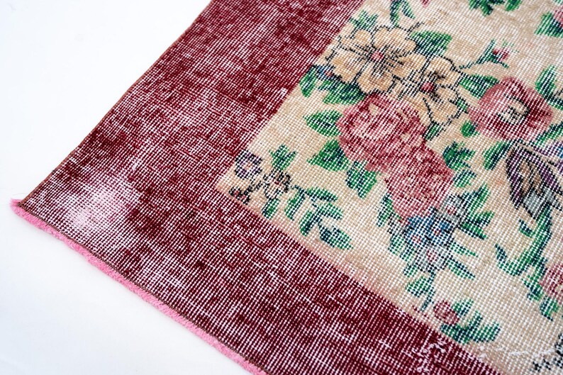 Pode incluir: Close-up de um tapete vintage com um padr&atilde;o floral. O tapete apresenta uma borda bord&ocirc; e um design central com flores rosa, verdes e azuis sobre um fundo bege. O tapete tem uma apar&ecirc;ncia desgastada e antiga.