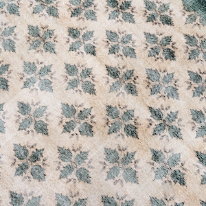 May include: A woven textile rug featuring a repeating pattern of dark teal four-leaf floral motifs arranged in a grid across a light beige background. The fabric displays a visible textured weave with a dark teal border along the top right edge.
