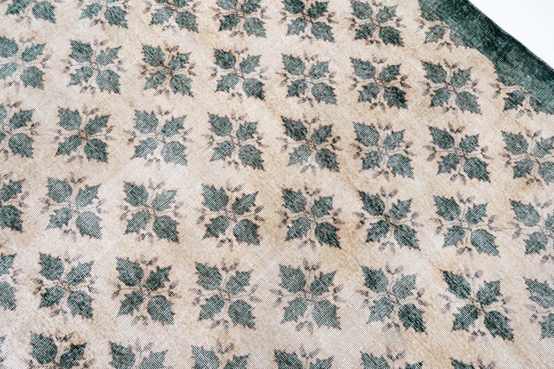 May include: A woven textile rug featuring a repeating pattern of dark teal four-leaf floral motifs arranged in a grid across a light beige background. The fabric displays a visible textured weave with a dark teal border along the top right edge.