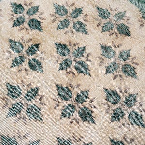 May include: A woven textile rug featuring a repeating pattern of dark teal four-leaf floral motifs set against a light beige background. The fabric displays a visible grid-like weave texture throughout, with a solid dark teal border along the edge.