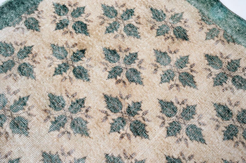 May include: A woven textile rug featuring a repeating pattern of dark teal four-leaf floral motifs set against a light beige background. The fabric displays a visible grid-like weave texture throughout, with a solid dark teal border along the edge.