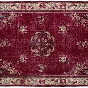 May include: A rectangular, distressed burgundy rug with a floral pattern. The rug features a central medallion and floral clusters, framed by a beige border with repeating geometric and floral motifs. The rug's design is symmetrical.