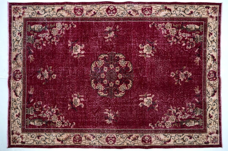 May include: A rectangular, distressed burgundy rug with a floral pattern. The rug features a central medallion and floral clusters, framed by a beige border with repeating geometric and floral motifs. The rug's design is symmetrical.
