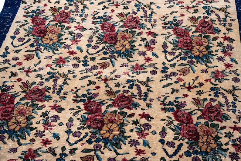 May include: Beige floral rug with a dark blue border. The rug features a repeating pattern of red, yellow, and purple flowers with green and blue leaves. The rug has a vintage, antique appearance.