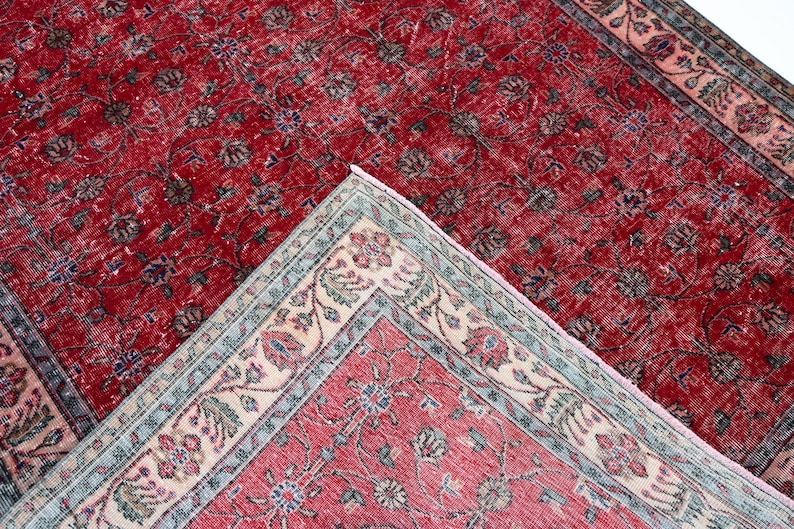 K&ouml;nnte beinhalten: Zwei antike rote Teppiche mit floralen Mustern. Ein Teppich ist vollst&auml;ndig sichtbar, w&auml;hrend der andere teilweise sichtbar ist, mit einer hochgeklappten Ecke, die einen detaillierten Rand mit cremefarbenen, blauen und rosa Akzenten zeigt. Die Teppiche haben einen Used-Look.