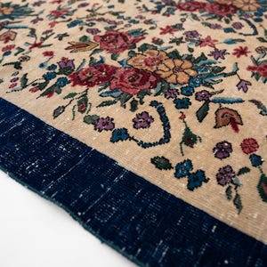 May include: Close-up of a beige rug with a floral pattern. The rug features a dark blue border and is adorned with colorful flowers in shades of red, blue, purple, and yellow. The rug's design is intricate and detailed.