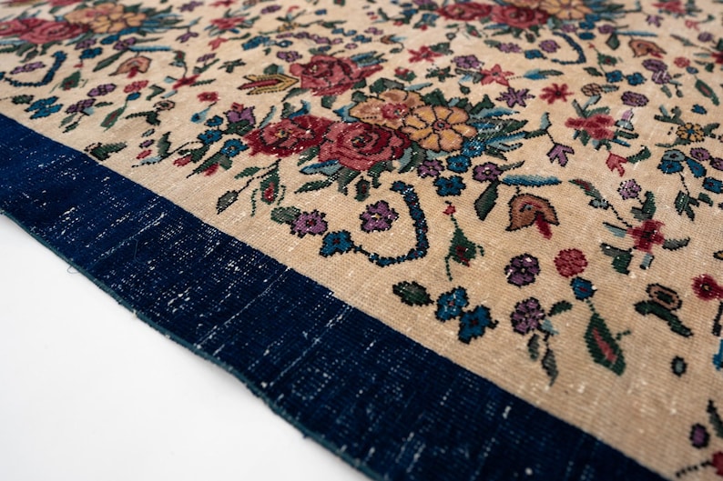 May include: Close-up of a beige rug with a floral pattern. The rug features a dark blue border and is adorned with colorful flowers in shades of red, blue, purple, and yellow. The rug's design is intricate and detailed.