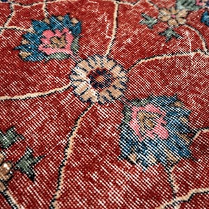May include: Close-up of a red, vintage-style rug with an intricate floral pattern. The design features stylized flowers in shades of blue, pink, and beige, set against a distressed red background. The rug's texture is visible, showcasing its woven construction.