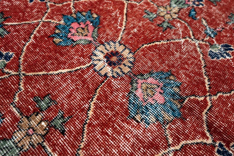 May include: Close-up of a red, vintage-style rug with an intricate floral pattern. The design features stylized flowers in shades of blue, pink, and beige, set against a distressed red background. The rug's texture is visible, showcasing its woven construction.