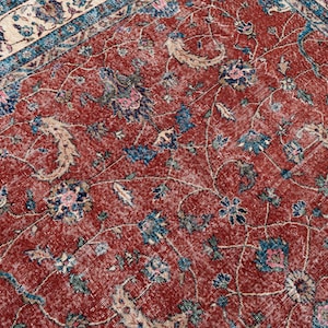 May include: Close-up of a distressed, vintage rug with a rich, red background. The rug features an intricate floral pattern in shades of blue, pink, and cream. The design includes stylized flowers and scrolling vines, creating a classic, aged aesthetic.