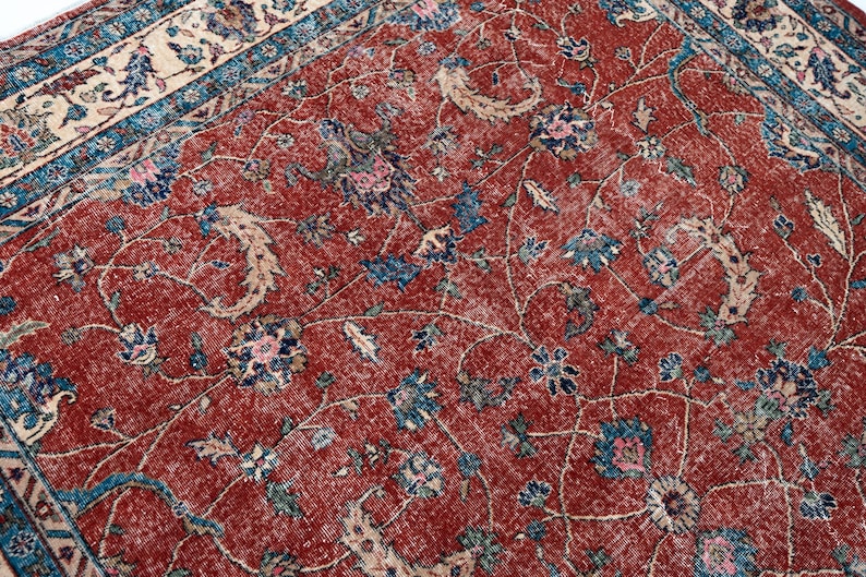 May include: Close-up of a distressed, vintage rug with a rich, red background. The rug features an intricate floral pattern in shades of blue, pink, and cream. The design includes stylized flowers and scrolling vines, creating a classic, aged aesthetic.
