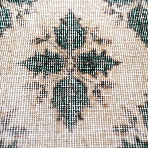 May include: A woven textile featuring a repeating pattern of dark green, leaf-shaped motifs arranged in a symmetrical grid. The background consists of a light beige, textured fabric with a visible grid-like weave structure throughout the surface.