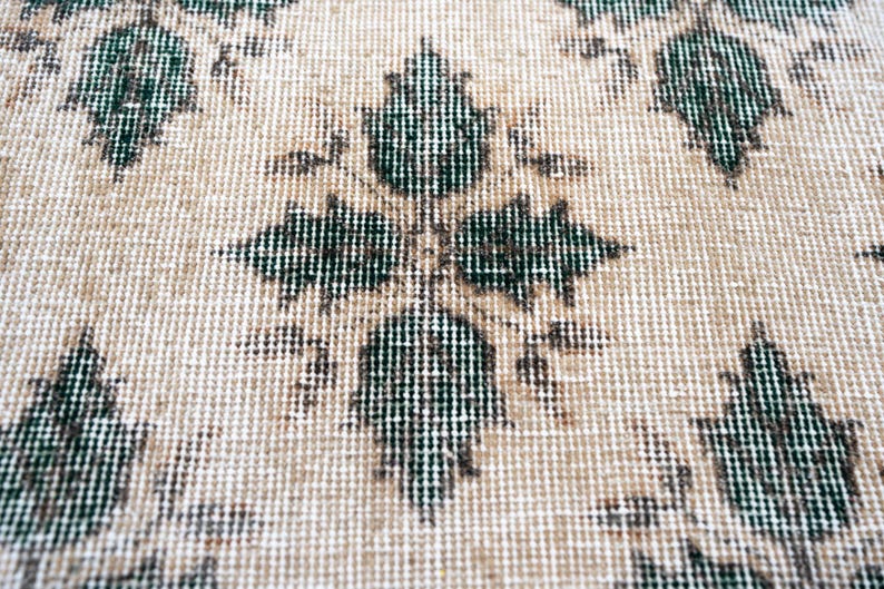 May include: A woven textile featuring a repeating pattern of dark green, leaf-shaped motifs arranged in a symmetrical grid. The background consists of a light beige, textured fabric with a visible grid-like weave structure throughout the surface.