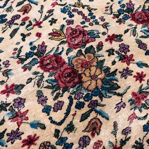 May include: Close-up of a beige rug with a detailed floral pattern. The rug features a variety of flowers in shades of red, purple, blue, and yellow, set against a neutral background. The design is intricate and appears to be vintage.