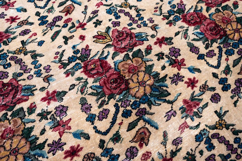 May include: Close-up of a beige rug with a detailed floral pattern. The rug features a variety of flowers in shades of red, purple, blue, and yellow, set against a neutral background. The design is intricate and appears to be vintage.