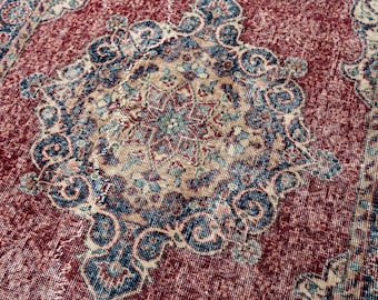 May include: Close-up of a distressed, vintage rug with a rich, burgundy background. The rug features a central medallion design in beige, teal, and navy blue, with intricate floral and geometric patterns. The rug's edges are also detailed.