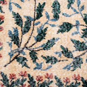 May include: Close-up of a beige rug with a floral pattern. The design features green and blue leaves with red flowers along the border. The rug has a textured appearance.