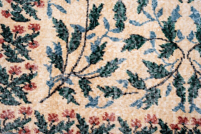 May include: Close-up of a beige rug with a floral pattern. The design features green and blue leaves with red flowers along the border. The rug has a textured appearance.