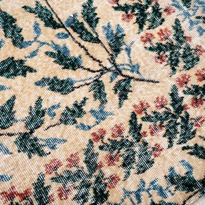 May include: Close-up of a beige rug with a floral pattern. The design features green leaves, blue accents, and red flowers. The rug has a distressed, vintage look, with a soft, textured surface.