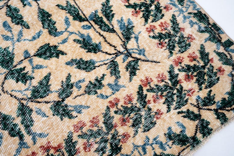 May include: Close-up of a beige rug with a floral pattern. The design features green leaves, blue accents, and red flowers. The rug has a distressed, vintage look, with a soft, textured surface.