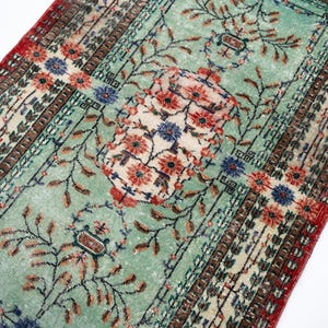 May include: An antique rug with a distressed, faded appearance. The rug features a central floral medallion in cream, red, and blue, set against a light green field. The borders are red and brown, with intricate floral and geometric patterns.