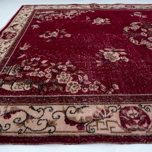 May include: A rectangular, vintage-style rug with a deep red field and a beige border. The rug features floral patterns in beige and brown, with a central medallion design. The border has intricate scrollwork and geometric elements.