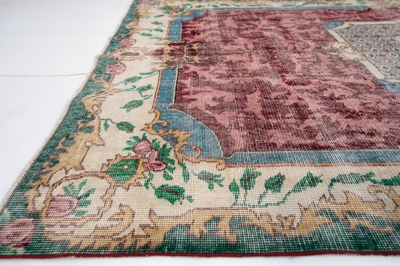 May include: An antique rug with a distressed, vintage appearance. The rug features a central maroon field with a floral pattern, framed by a cream border with green, blue, and gold accents. The rug's design includes floral and botanical motifs.