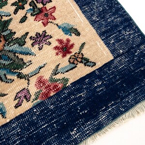 May include: Close-up of a vintage rug with a floral pattern. The rug features a cream-colored central field with colorful flowers in shades of pink, yellow, and blue. A dark blue border frames the design, and the rug has a fringed edge.