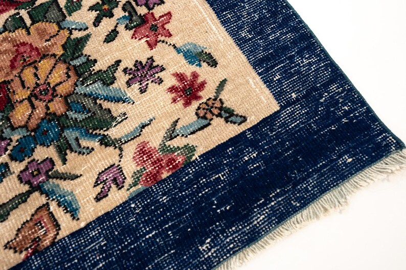 May include: Close-up of a vintage rug with a floral pattern. The rug features a cream-colored central field with colorful flowers in shades of pink, yellow, and blue. A dark blue border frames the design, and the rug has a fringed edge.