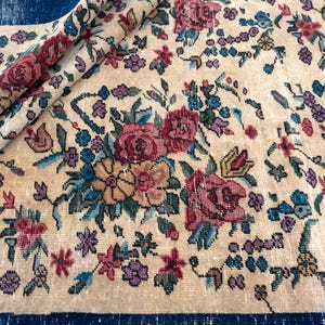 May include: Cream-colored vintage rug with a floral pattern. The rug features a repeating design of roses, other flowers, and foliage in shades of red, pink, blue, and yellow. The rug is partially rolled up, revealing a dark blue border.