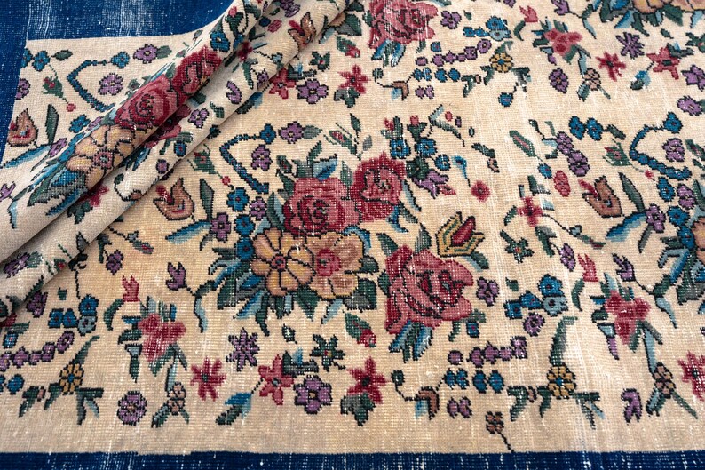 May include: Cream-colored vintage rug with a floral pattern. The rug features a repeating design of roses, other flowers, and foliage in shades of red, pink, blue, and yellow. The rug is partially rolled up, revealing a dark blue border.