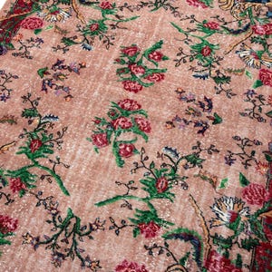 May include: A rectangular area rug featuring a dusty pink background with a repeating floral pattern in shades of red, green, and dark blue. The rug has a wide, dark red border decorated with intricate floral motifs and scrolling vine details.