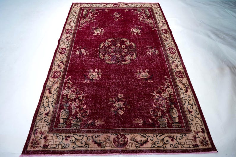 May include: A rectangular, vintage-style rug with a deep red field and a beige border. The rug features floral patterns and a central medallion design. The border has intricate scrollwork and floral motifs. The rug's design suggests a classic, antique aesthetic.