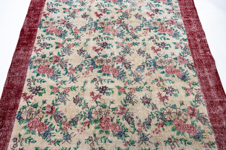 Pode incluir: Um tapete floral vintage com fundo creme e um padr&atilde;o de flores rosa, azuis e roxas. O tapete tem uma borda vermelha escura. Adequado para salas de estar e quartos. Mede aproximadamente 200 cm x 300 cm.