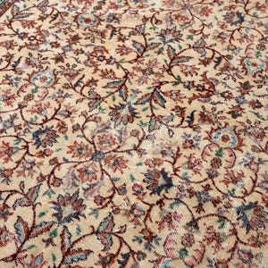 May include: Close-up of a beige rug with an intricate floral pattern in shades of red, blue, and green. The rug has a detailed border with a teal and brown design. The overall texture appears soft and aged.