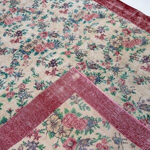 Pode incluir: Um tapete com padr&atilde;o floral com fundo creme e borda vermelha escura. O tapete apresenta um padr&atilde;o repetido de flores rosa, azuis e roxas com folhas verdes. O tapete retangular parece ser feito de material tecido.