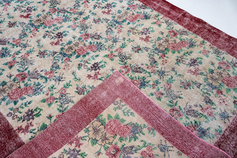 Pode incluir: Um tapete com padr&atilde;o floral com fundo creme e borda vermelha escura. O tapete apresenta um padr&atilde;o repetido de flores rosa, azuis e roxas com folhas verdes. O tapete retangular parece ser feito de material tecido.