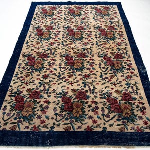 May include: Rectangular beige rug with a dark blue border. The rug features a repeating floral pattern in shades of red, pink, yellow, and blue. The rug is likely made of woven material and is suitable for home decor.