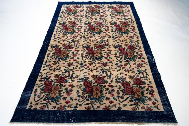 May include: Rectangular beige rug with a dark blue border. The rug features a repeating floral pattern in shades of red, pink, yellow, and blue. The rug is likely made of woven material and is suitable for home decor.