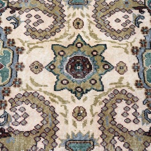 May include: A woven rug featuring a repeating pattern of large, stylized floral motifs and geometric shapes in shades of sage green, dusty blue, cream, and deep maroon. The intricate design includes central star-like medallions and curved paisley-inspired elements arranged across a light-colored background.