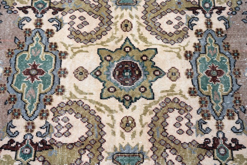May include: A woven rug featuring a repeating pattern of large, stylized floral motifs and geometric shapes in shades of sage green, dusty blue, cream, and deep maroon. The intricate design includes central star-like medallions and curved paisley-inspired elements arranged across a light-colored background.