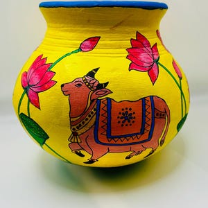 May include: A hand-painted yellow ceramic pot with a blue rim, featuring a pink cow and lotus flowers. The cow has a decorative saddle and is surrounded by green stems and pink flowers. The pot is a decorative item.