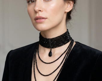 Puede incluir: Un collar tipo gargantilla de cuentas negras de varias capas con un colgante central en forma de lágrima. La joya consta de una banda ancha de cuentas ajustada al cuello con tres hilos de cuentas largos y delicados que caen hacia abajo.